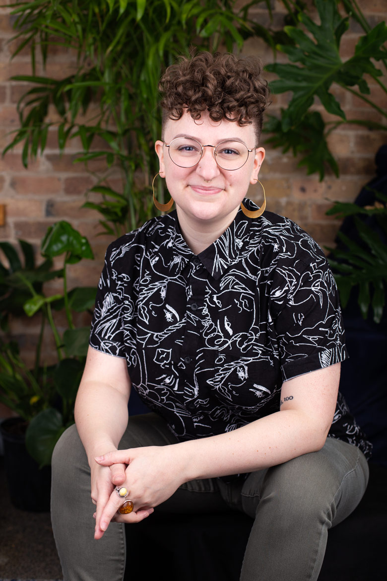 Jordan, a white agender Woman with dark blonde hair - curly and long on the top and shaved short on the sides - smiles at the camera. They are wearing a black short sleeve button up shirt with white line drawings of faces, gold earrings, and round, gold-rimmed glasses.