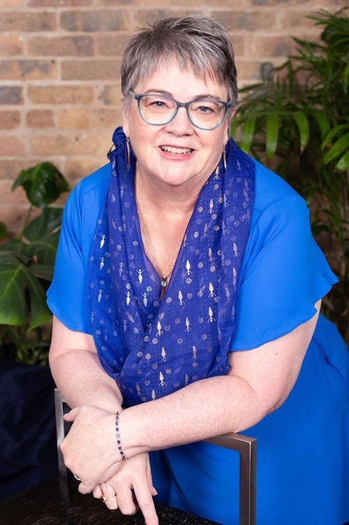 Betty Guilfoile, a white cisgender older woman with short gray hair and blue glasses, smiles at the camera while wearing a sky blue blouse with a darker blue scarf and gold earings.