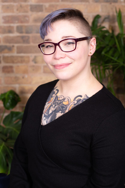 Gia Scalise, a white genderqueer person, sits angled to the left and smiles close-lipped. They have an undercut with short, lavender-colored hair falling over their forehead. She is wearing red, rectangle-frame glasses and a black, v-neck shirt that shows an animal-themed chest tattoo.