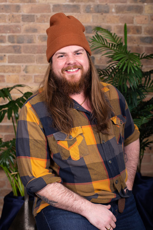 John, a white non binary person with long light brown hair and a brown with hints of red beard smiles at the camera. they are wearing a brown beanie and a blue and yellow flannel shirt. They have their left eyebrow pierced and turquoise plugs in their ear lobes.