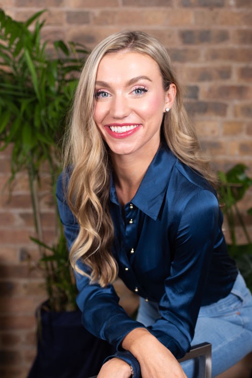 Sophia, a white ciswoman in her late twenties, stands behind a chair and leans forward with her arms folded over the back of the chair in a friendly, casual, way. Sophia has a big, toothy smile, rosy cheeks, blue eyes, and long, blonde, wavy hair. She is of medium height and slight build. She wears a deep blue silk blouse with brass buttons down the front and blue jeans.