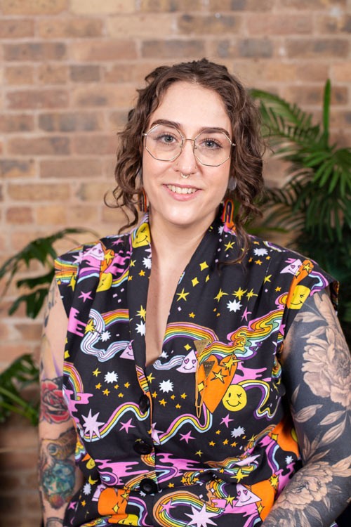 Alex, a white non-binary woman with brown curly hair framing their smiling face wears a septum ring, hexagonal gold framed glasses, and raibow earrings that peak out from behind her hair. Her sleeveless buttoned up jumpsuit has a bright raindow, heart, and smiley faced pattern covering its black background. Her body is slightly angled so her left arm is in the foreground displaying thier black and grey tattoo sleeve. In a background you can see that her right arm has multiple colored tattoos covering it as well, including one of a cuttlefish.