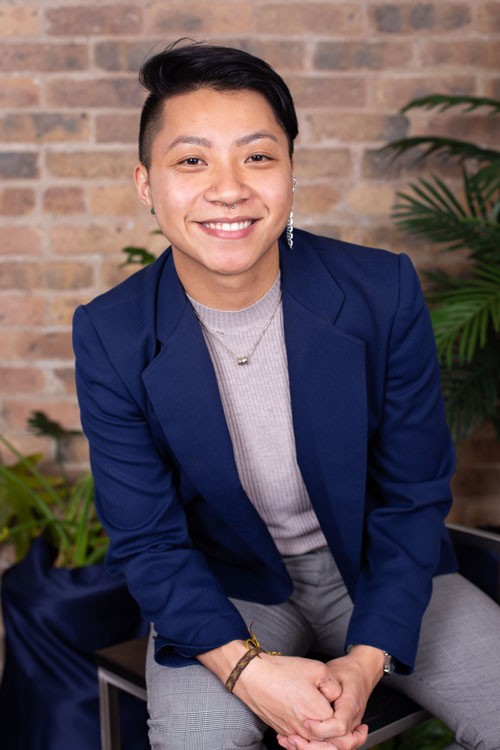 Eros, a light skinned Chinese-American, genderqueer, transmasculine person with short black hair brushed to the left and shaved short on the sides - leaning forward and smiling at the camera. They are wearing a beige turtle neck with a navy blue blazer, wearing a gold necklance, one silver dangling earing on their upper left ear, and a threaded brown and yellow bracelet on their right wrist. Behind them is a blurred red brick wall and palm plant.