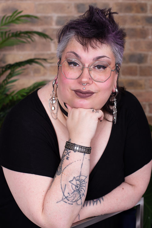 Vesryn Grey, a white trans non-binary queer person, is leaning forward on a chair, right hand under their chin, smirking at the camera. Behind them is a blurred red brick wall and palm plant. They have a modern mullet, their hair being black and silver, with touches of purple in it. They are wearing silver hexagon glasses, purple eye makeup, and purple-ish lipstick. They have a nostril, septum, and philtrum piercings. They have stretched lobes wherein they sport hanging bejeweled bee earrings. On their right wrist is a leather bracelet and they also wear a matching leather collar. They have on a v-necked black jumpsuit. On their arms is a multitude of tattoos.