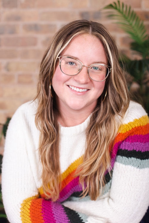 Cat Kliegle, a white person with long blonde hair, leaning forward towards the camera with their arms crossed. They are wearing a white sweater with a rainbow stripe diagonally across the front of the sweater. She is also wearing gold-framed glasses and gold hoop earrings.