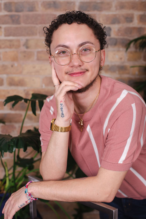Jae Avilez, wearing a primarily pink and white striped short sleeve sweater, leans over a tall chair with their right hand under their chin. They're smiling at the camera, wearing round gold glasses and in front of a brick wall and tall green plants.