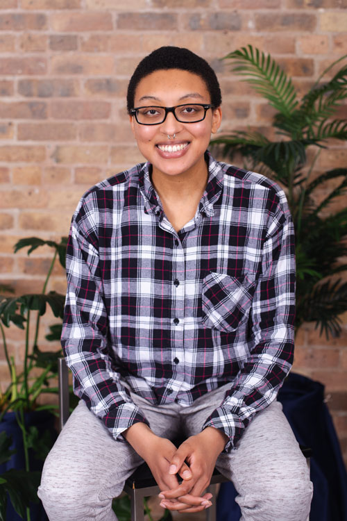 Maya Vadell, wearing a black and white plaid shirt and leaning slightly forward on a stool smiles at the camera.