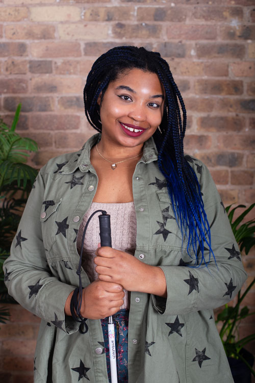 Janay, a medium-toned black woman smiles at the camera while holding a long white cane.