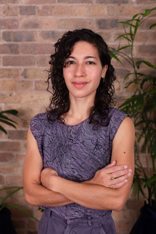 Sarah Schaffer smiles with arms gently crossed. She is a mixed Jewish and Southeast Asian person with light brown skin and dark curly hair in her thirties.